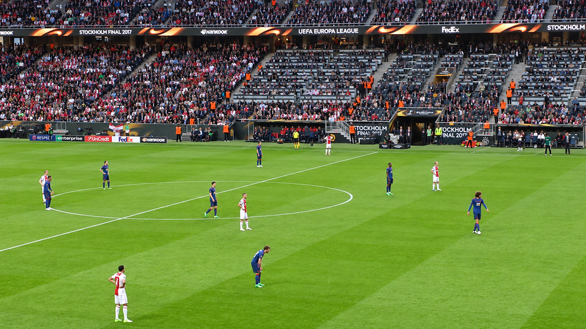 Soccer players stand on a green field before kickoff at a stadium packed with fans. The scoreboard displays “STOCKHOLM FINAL 2017” and “UEFA EUROPA LEAGUE.”
