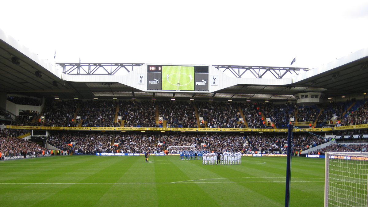 A soccer stadium with a crowd in the stands. Two teams stand in lines on the field, possibly before kickoff. The scoreboard and a large screen are visible above the stands.