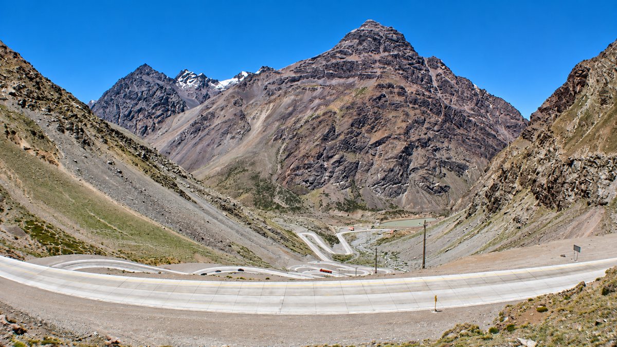 Winding mountain road with sharp curves, surrounded by rocky slopes and peaks under a clear blue sky.