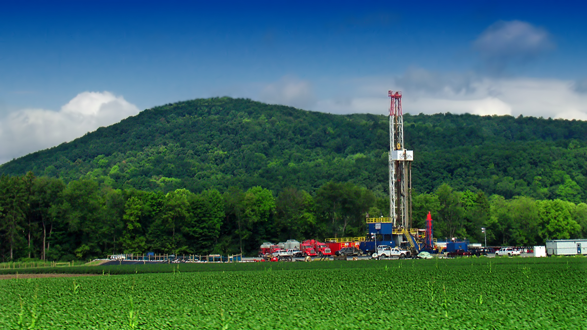 A drilling rig stands in a green field with vehicles and equipment nearby, set against a backdrop of dense trees and a forested hill under a blue, partly cloudy sky.