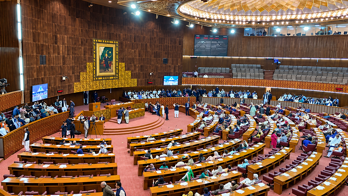 A large parliamentary chamber with rows of curved desks, many seated people, some standing, and a large portrait on the wall. The ceiling is ornate, and electronic boards display information.