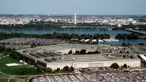 Aerial view of the Pentagon building surrounded by parking lots, with the Washington Monument and cityscape visible in the background.