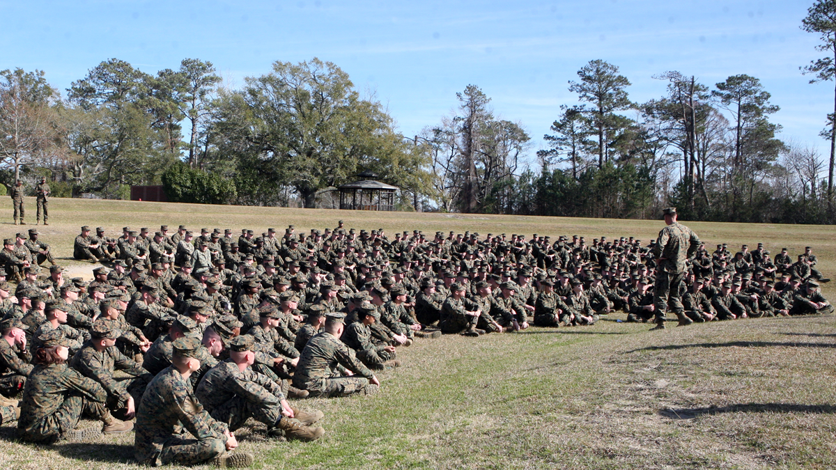 A large group of people in military uniforms sit on grass, listening to a person standing in front of them outdoors, surrounded by trees under a blue sky.