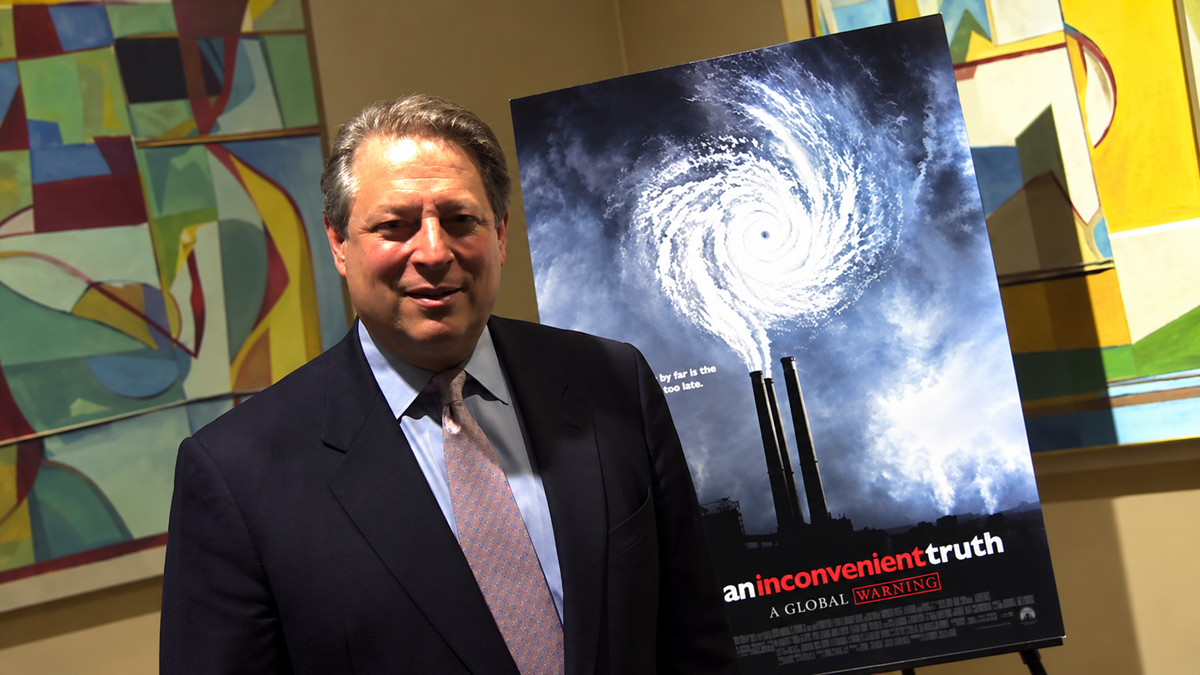 A man in a suit stands next to a poster for the film “An Inconvenient Truth,” featuring a swirling storm above factory smokestacks. Abstract paintings are visible on the wall behind him.