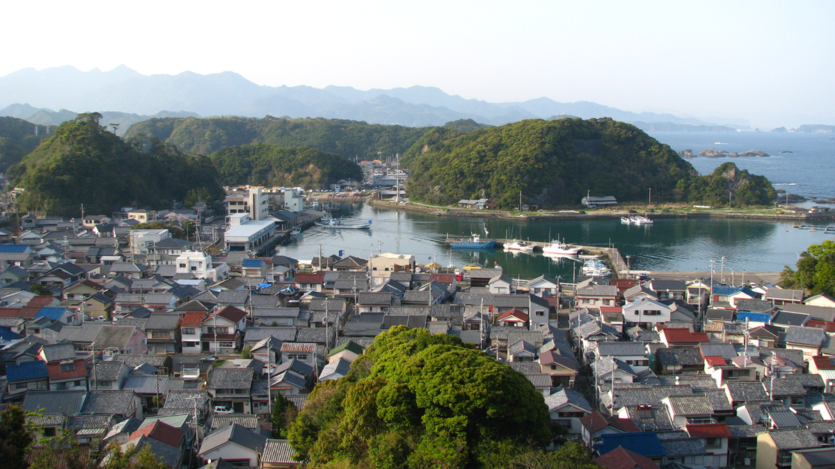 A coastal Japanese town with tightly packed houses, a harbor with boats, green hills surrounding the water, and distant mountains under a clear sky.