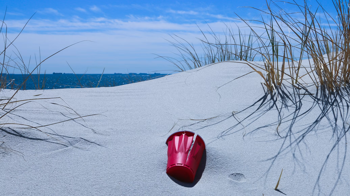 A red plastic cup lies on white sand near tall grass, with the ocean and distant ships visible under a blue sky.