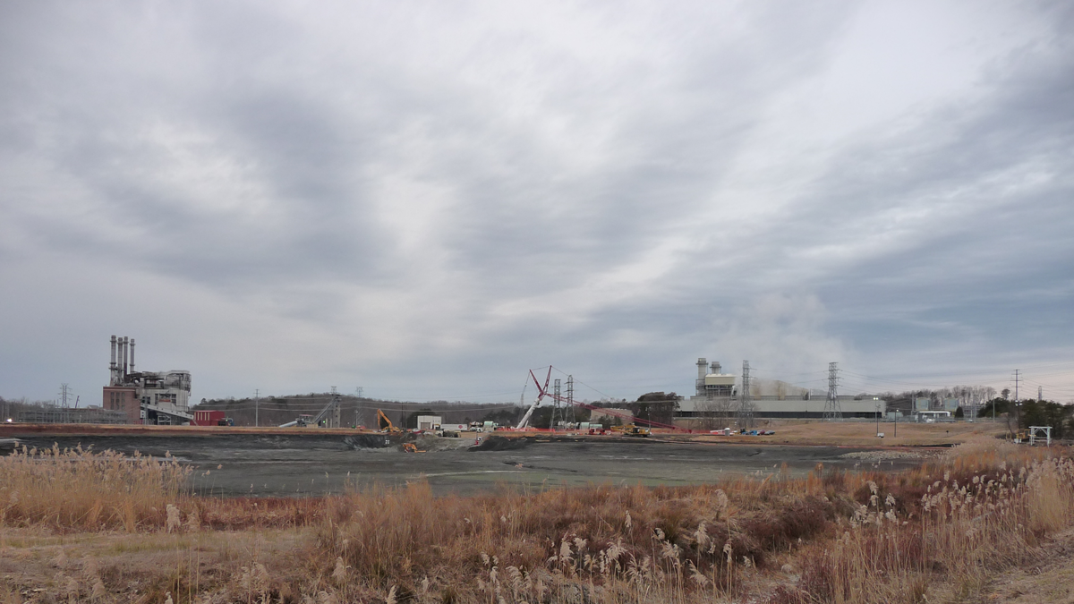 A cloudy sky over an industrial area with smokestacks, cranes, construction equipment, and dry grass in the foreground.
