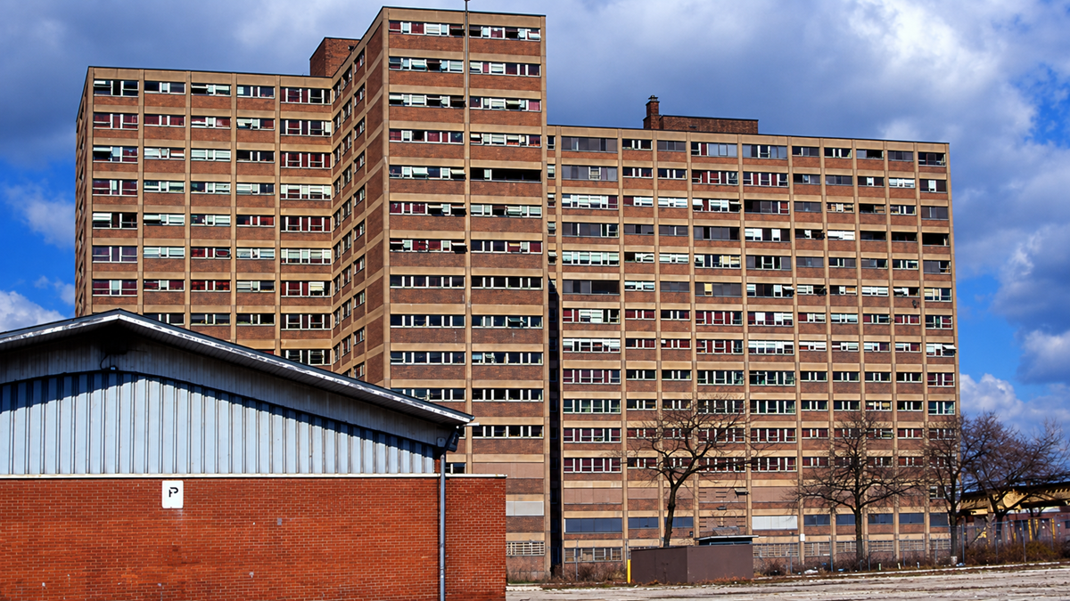 A tall, old brick apartment building with many windows, some broken, stands behind a shorter industrial building and leafless trees under a partly cloudy sky.