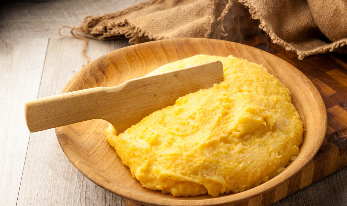 Food. A wooden bowl filled with creamy yellow polenta and a wooden spatula, placed on a rustic wooden table with burlap fabric in the background.