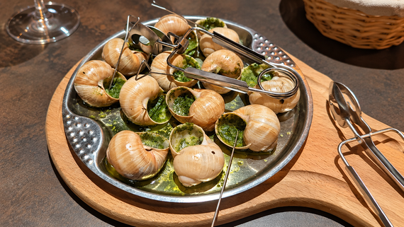 A metal tray holds cooked escargot with green herb butter, served with tongs and picks on a wooden board. A basket is visible in the background.