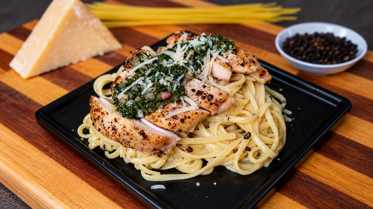 A square black plate with fettuccine pasta topped with sliced grilled chicken and pesto, garnished with grated cheese. In the background are a wedge of cheese, dry spaghetti, and a bowl of black peppercorns.