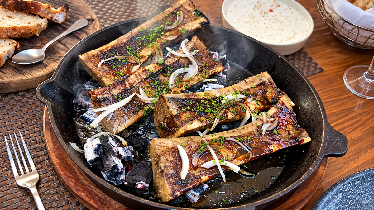 Four roasted bone marrow halves garnished with herbs and onion slices are served in a cast iron skillet, with a side of bread, dipping sauce, and utensils on a wooden table.