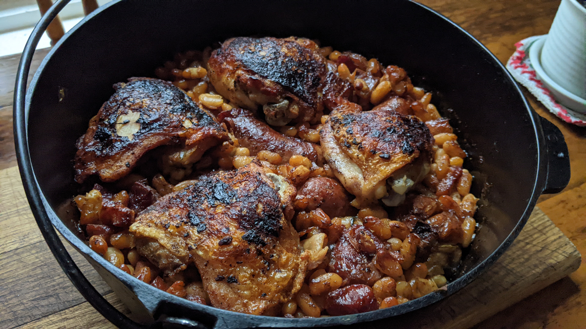 A cast iron pot filled with browned chicken thighs on top of baked beans, resting on a wooden surface.