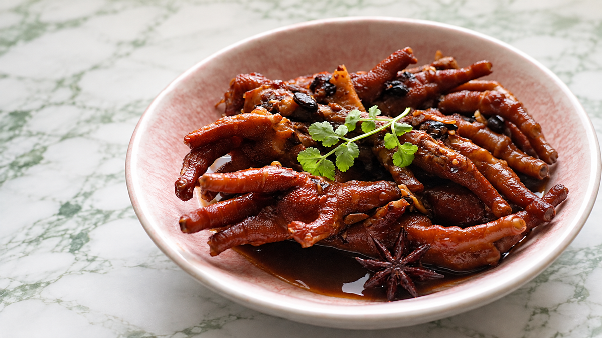 A bowl of braised chicken feet garnished with cilantro and star anise, served in a brown sauce on a pink-rimmed plate, placed on a white and green patterned surface.