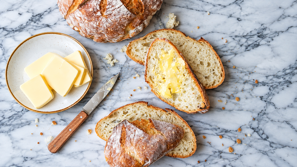 Sliced crusty bread on a marble surface, with one slice buttered. Nearby are a plate with butter slices and a butter knife.