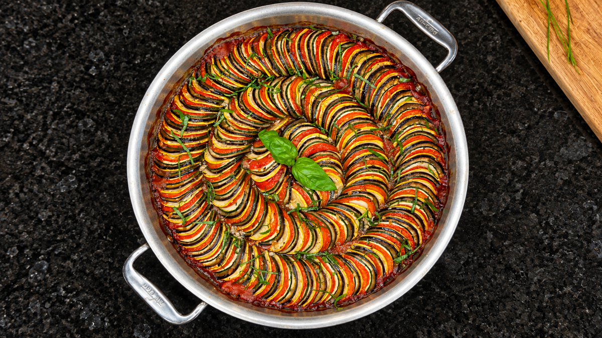 A round metal pan filled with neatly arranged, spiral slices of zucchini, tomato, and eggplant, garnished with two fresh basil leaves, sits on a black countertop.