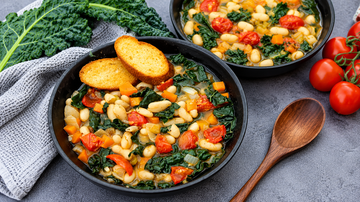 Two bowls of vegetable soup with white beans, kale, tomatoes, and carrots, topped with toasted bread slices. A wooden spoon, fresh kale, and tomatoes are beside the bowls on a gray surface.