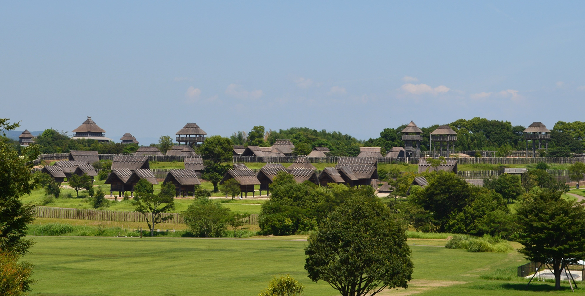 A historical Japanese village with wooden buildings and watchtowers surrounded by trees and green grass under a clear blue sky.