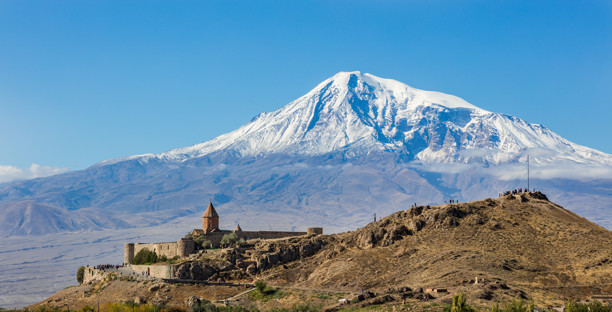 Snow-capped Mount Ararat rises behind Khor Virap Monastery and a sunlit, rocky hill with people standing on top, under a clear blue sky.