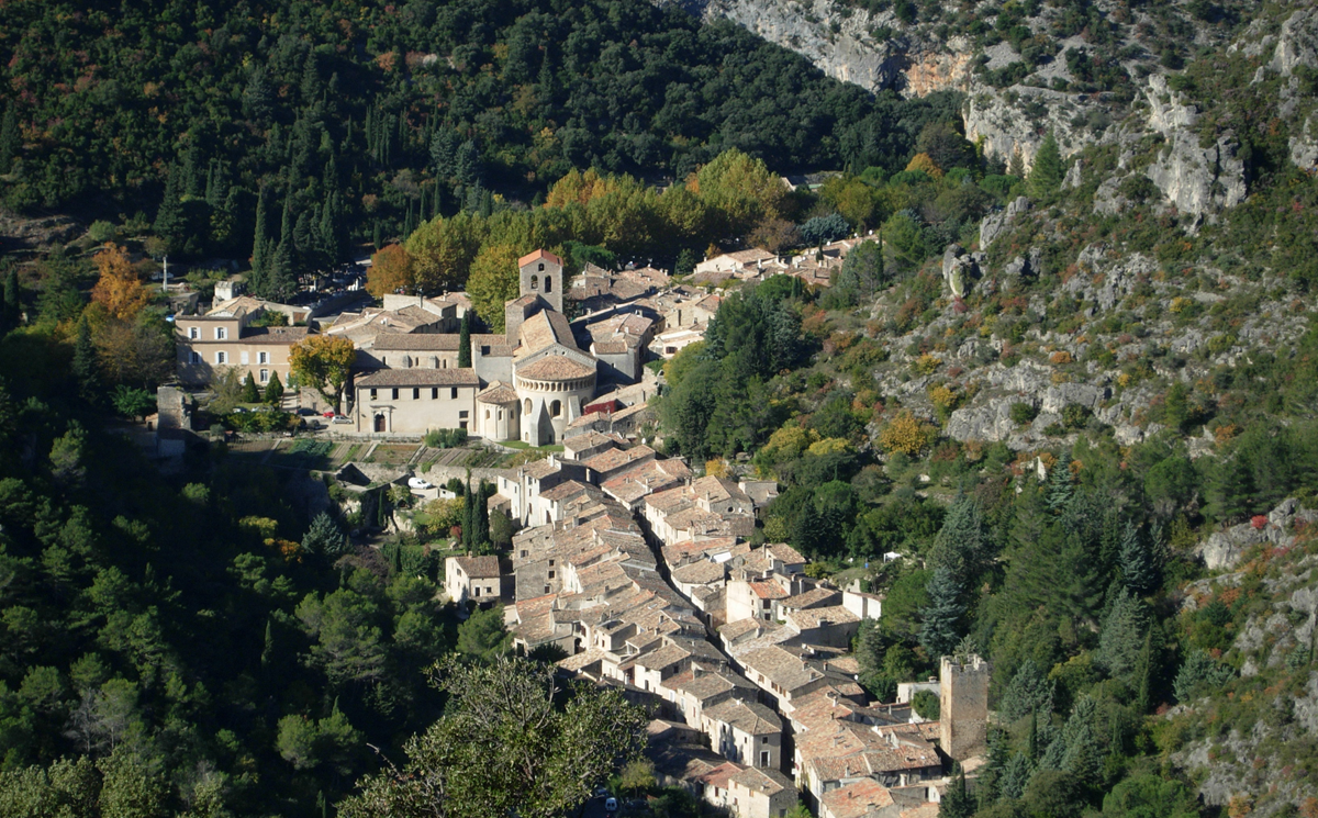 Aerial view of a small, historic village nestled in a green, forested valley, featuring stone buildings with tiled roofs and a prominent church surrounded by cliffs.