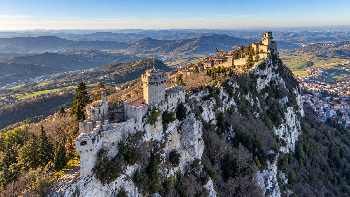 Stone towers and fortress walls sit atop a rocky hill surrounded by lush forests and a distant view of rolling hills and valleys under a clear blue sky.