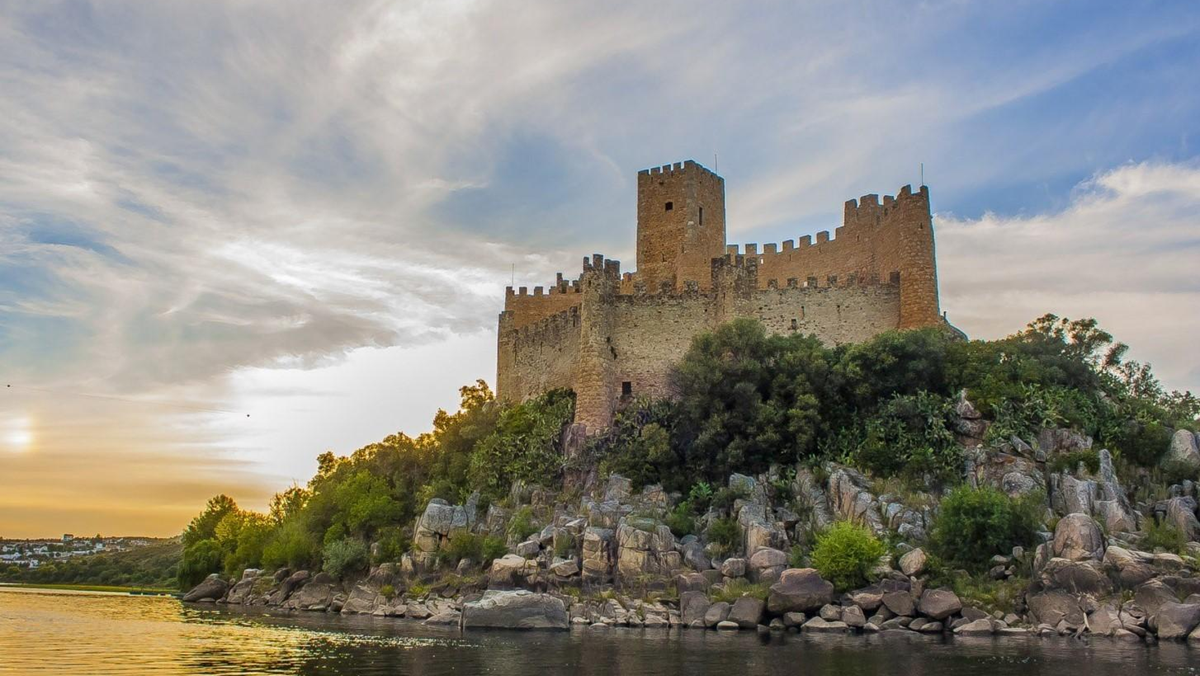 A stone castle sits atop a rocky, tree-covered hill beside a calm river at sunset, with partly cloudy skies in the background.