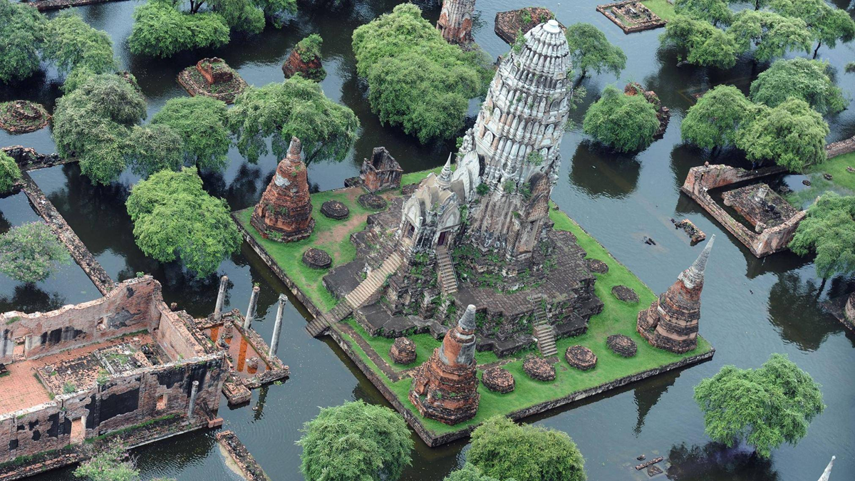 Aerial view of ancient temple ruins surrounded by floodwaters and green trees, with some buildings partially submerged.