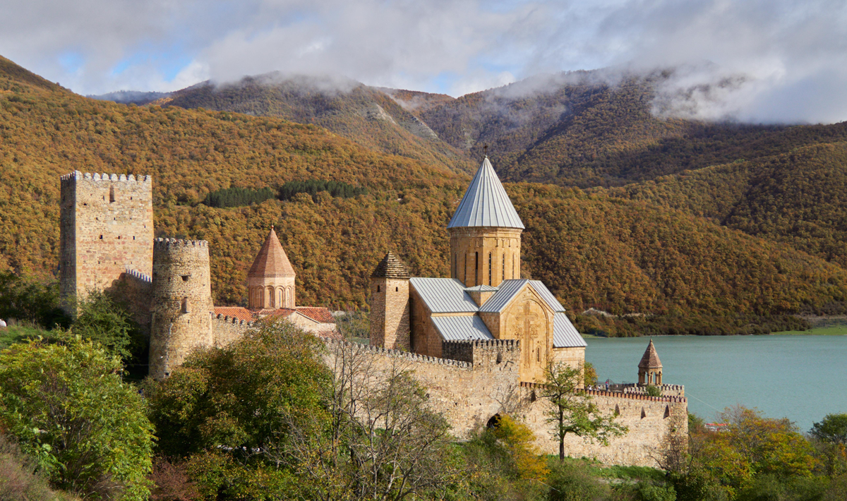 Ancient stone fortress with towers and a church sits by a turquoise lake, surrounded by forested mountains with autumn foliage under a partly cloudy sky.