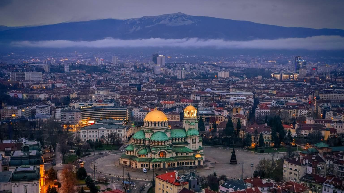 A cityscape at dusk featuring a large, green-domed cathedral in the foreground, surrounded by buildings, with mountains and a low-lying cloud in the background.