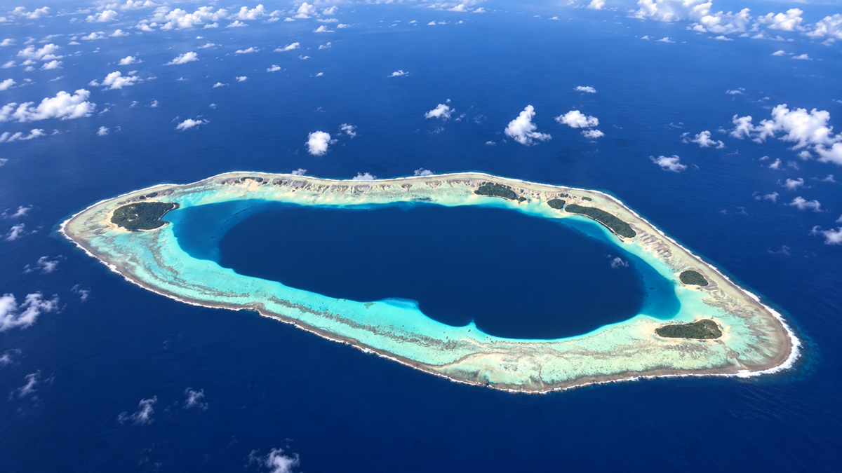 Aerial view of a circular coral atoll with a deep blue lagoon in the center, surrounded by turquoise shallow waters and small patches of green vegetation, set in the open ocean under a partly cloudy sky.