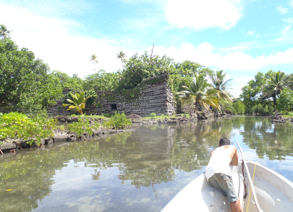 A person in a small boat approaches ancient stone ruins surrounded by lush greenery, palm trees, and calm water under a partly cloudy sky.
