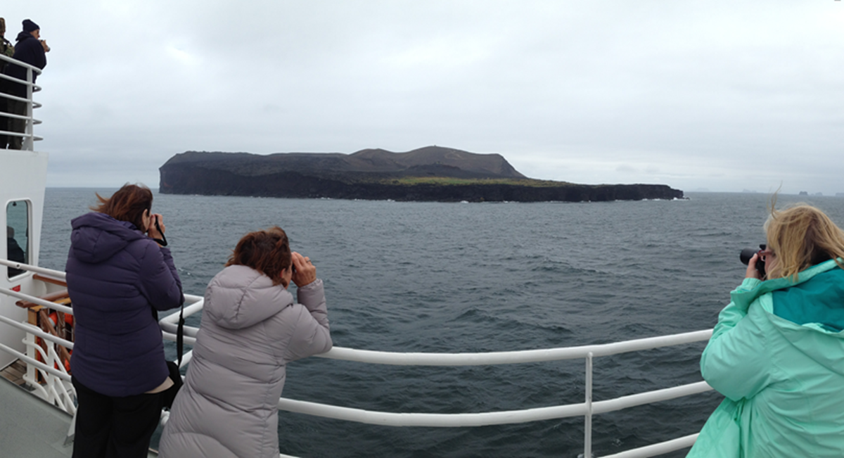Three people wearing winter jackets stand on a boat’s deck, looking at and photographing a rocky island with green patches across choppy grey water under an overcast sky.