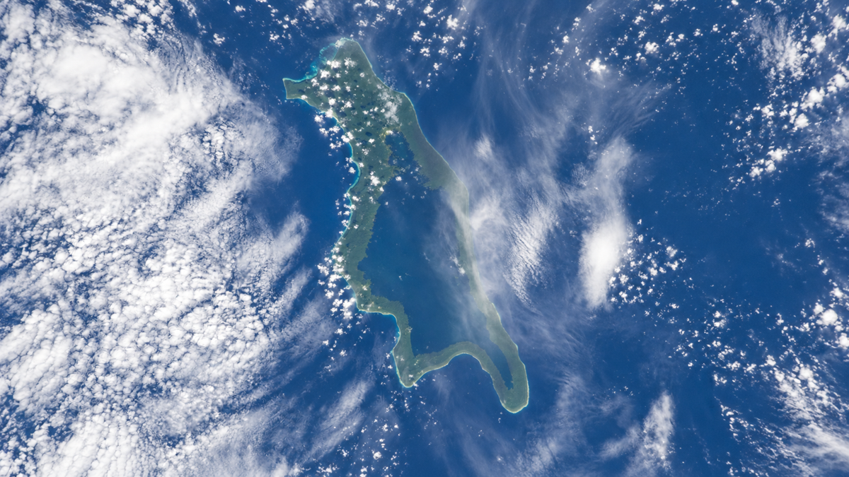 A satellite view of a green, irregularly shaped island surrounded by blue ocean and wispy white clouds scattered across the sky.