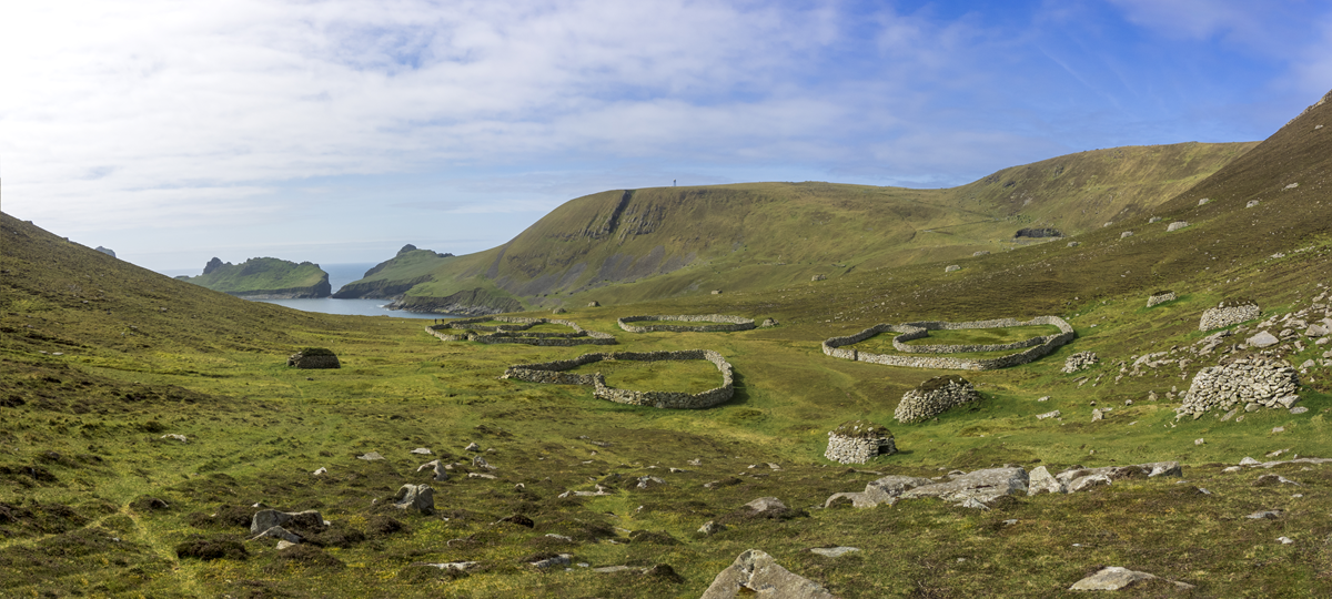 Ancient stone enclosures sit on a grassy, hilly landscape near the coastline under a partly cloudy sky, with cliffs and rocky outcrops in the distance.