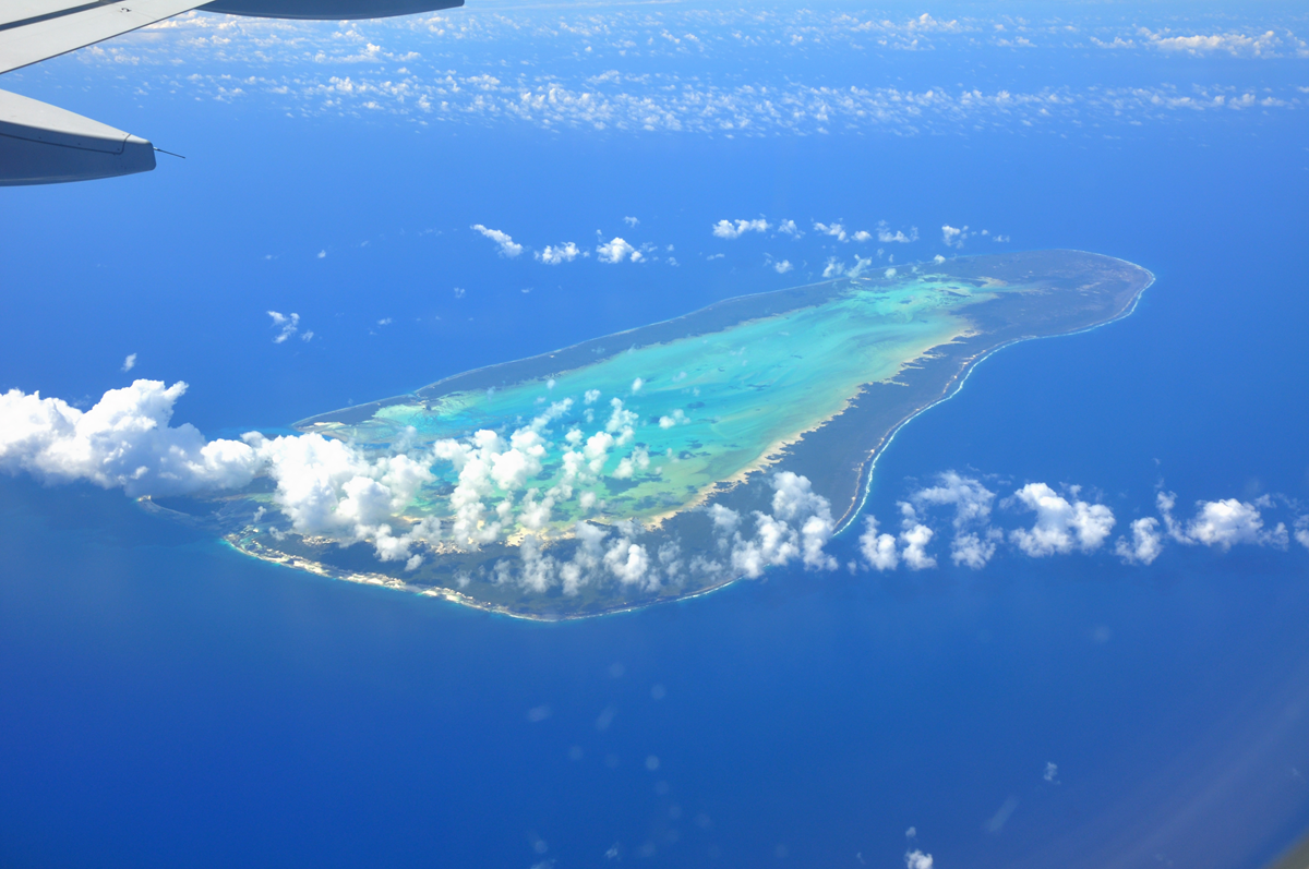 UNESCO Site. Aerial view of a tropical island surrounded by blue ocean, with scattered clouds overhead. Part of an airplane wing is visible at the top left of the image.