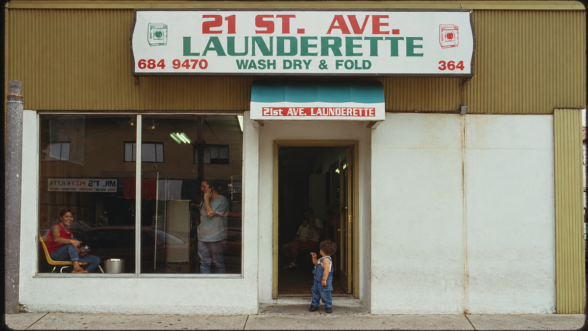 A child stands outside a laundromat with a “21 St. Ave. LAUNDERETTE” sign. Two people sit inside near large laundry machines. The storefront is plain with off-white walls.