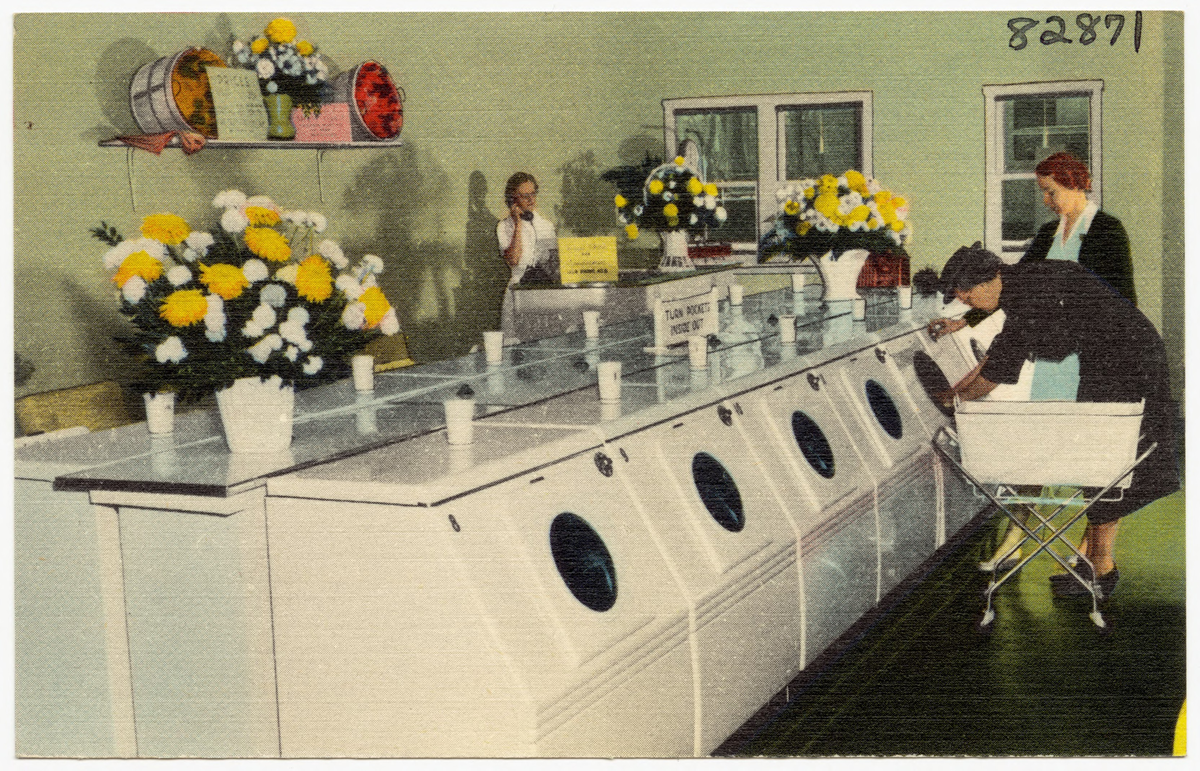 Women use front-loading washing machines in a bright laundromat decorated with large vases of yellow and white flowers on the machines and shelves. One woman stands by the counter, using a phone.