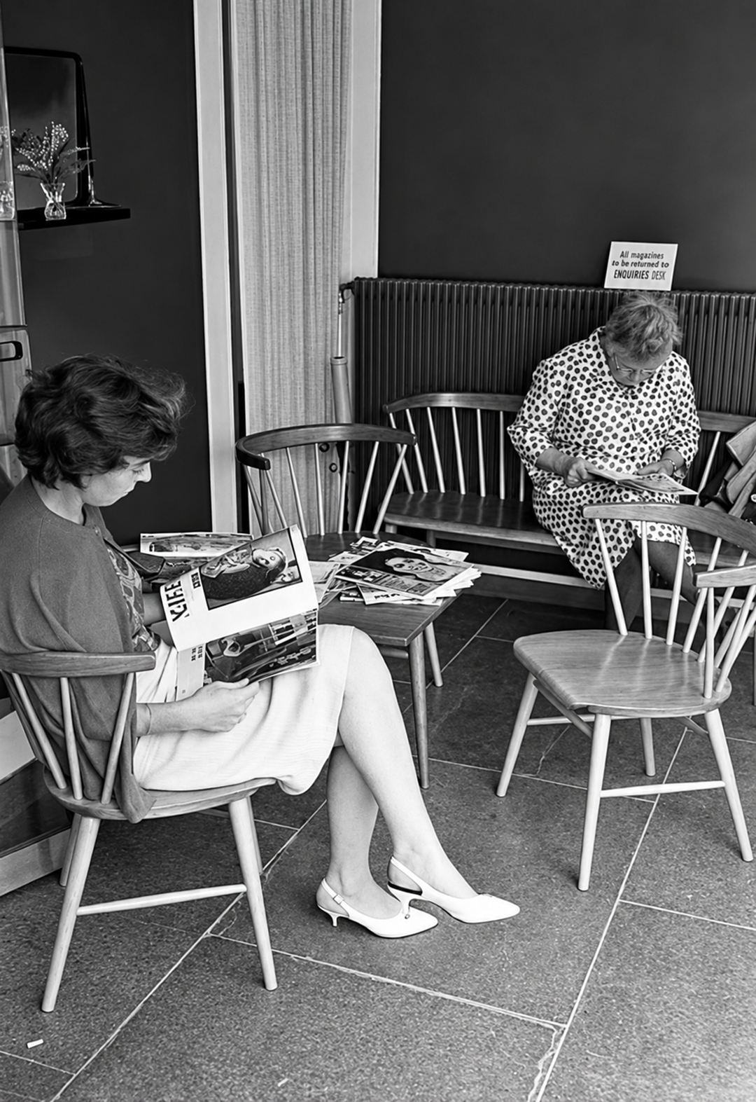 Two women sit in a waiting room; one reads a magazine, while the other knits. Wooden chairs, a small table, and a sign on the radiator are visible. The scene is black and white.