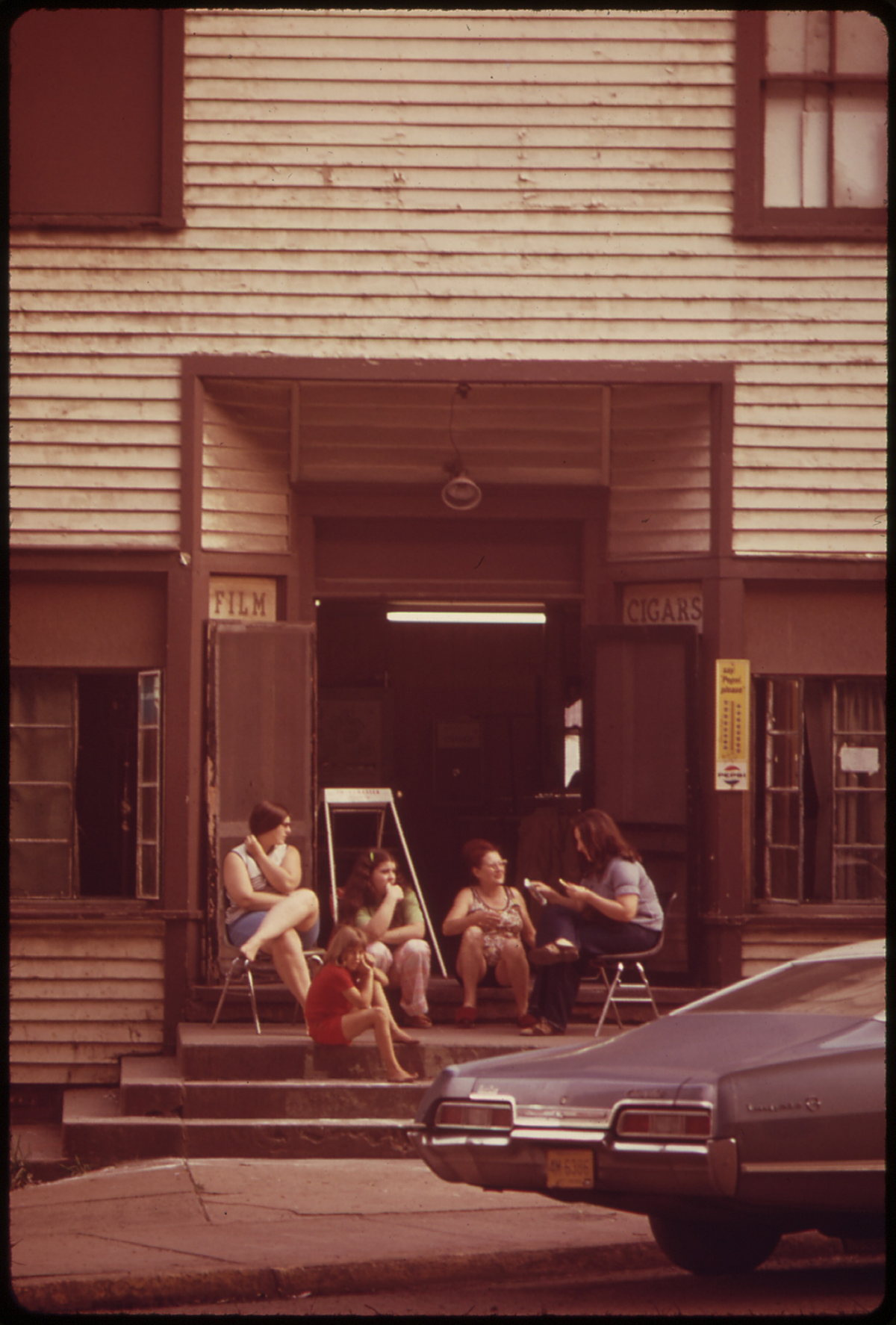 Four adults and a child sit on steps outside a worn, wooden building with signs for film and cigars. A blue car is parked in the foreground. The scene appears relaxed and casual.