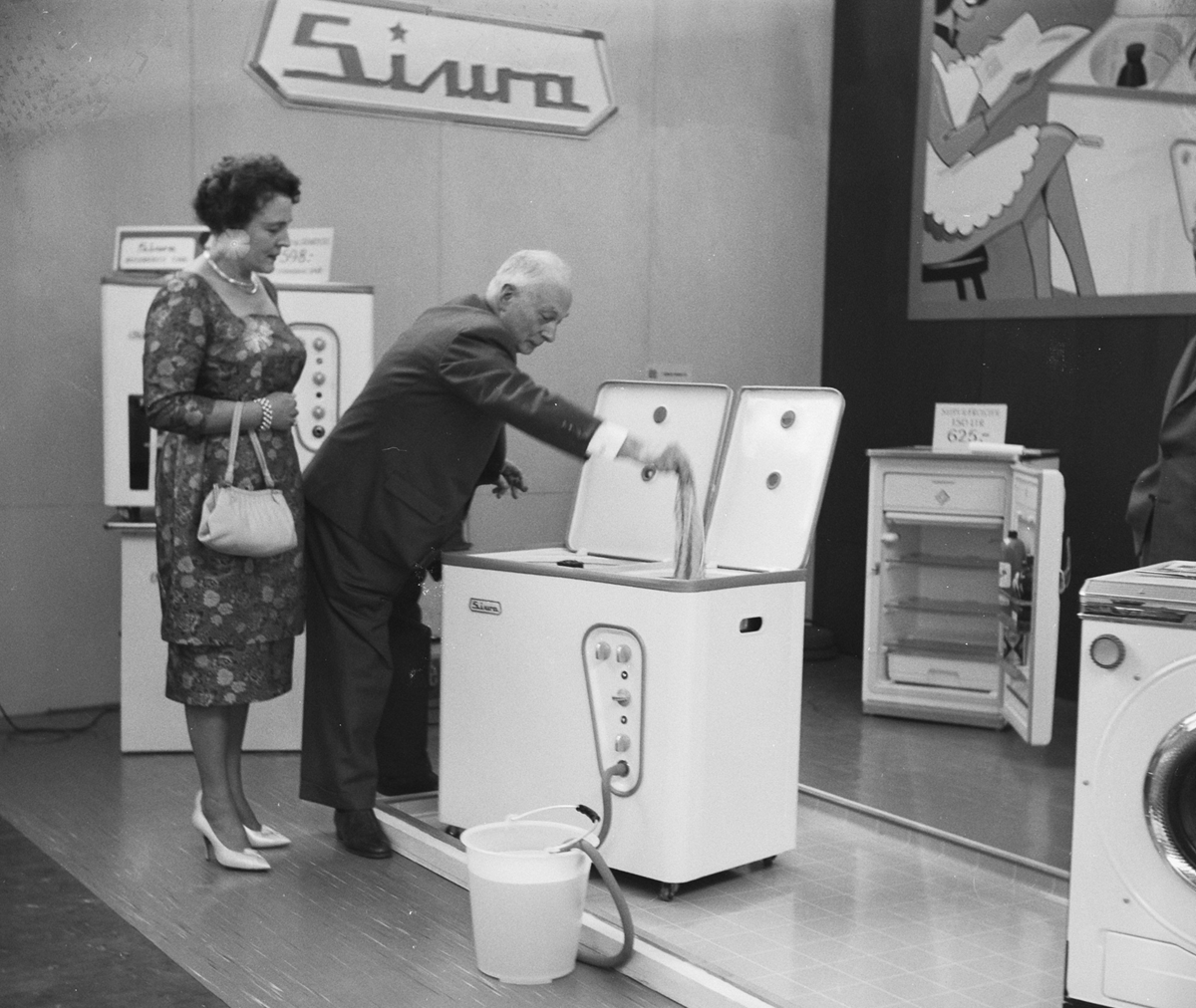 A man demonstrates a vintage washing machine by placing clothes inside, while a woman watches. They stand in an exhibition space with other appliances and product signs visible in the background.