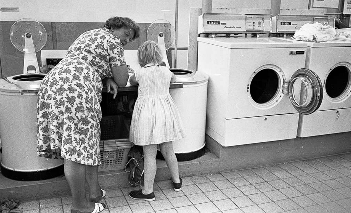 An older woman and a young girl look into a washing machine in a laundromat. The woman wears a floral dress; the girl wears a light dress and dark shoes. Nearby, empty dryers and folded laundry are visible.