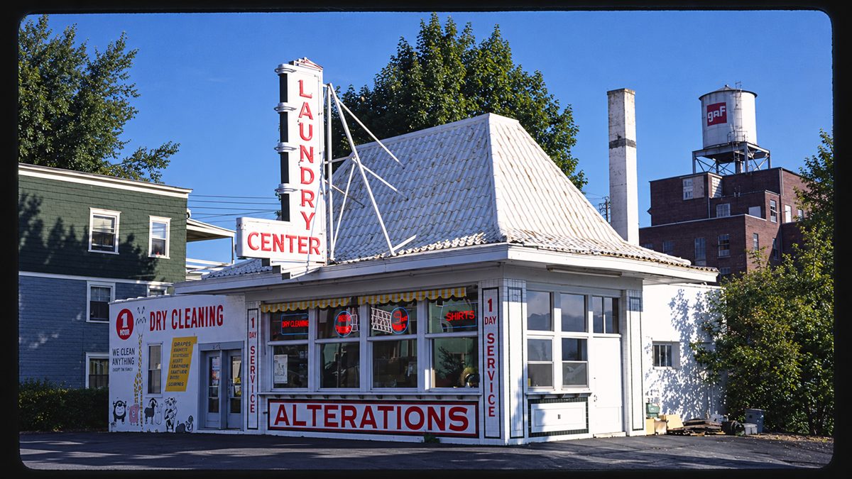 Small white building with a steep roof, large signs reading “Laundry Center” and “Alterations.” Neon signs and service windows are visible. Trees, a green house, and a water tower are in the background.