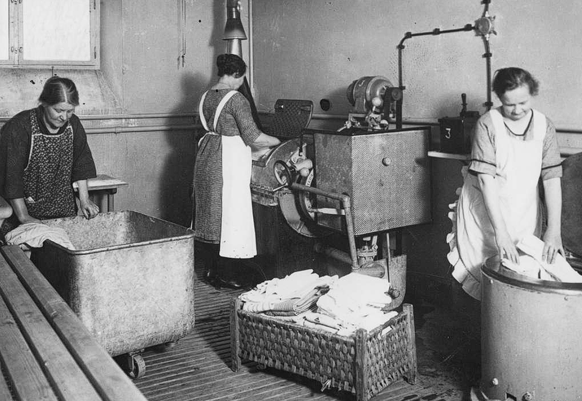 Three women in aprons work in an old-fashioned laundry room, washing and wringing clothes with large tubs and early washing machines.