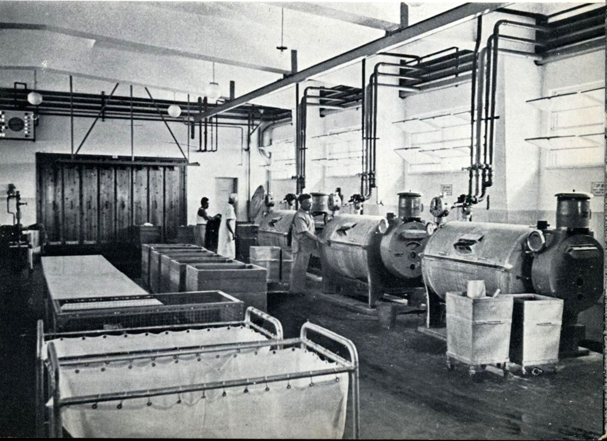 Black and white photo of a large laundry room with industrial washing machines, carts, baskets, and two people working near the machines. Pipes line the ceiling and walls, and light streams in through high windows.