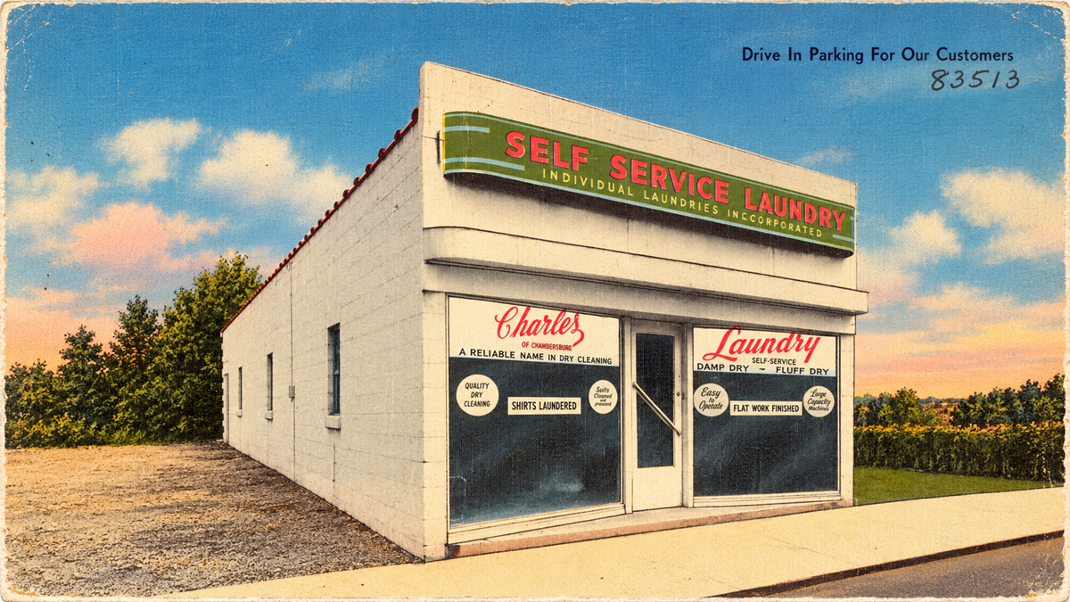 A vintage postcard shows a white building with a green “Self Service Laundry” sign. The windows advertise laundry and dry cleaning services. Trees and a blue sky are in the background.