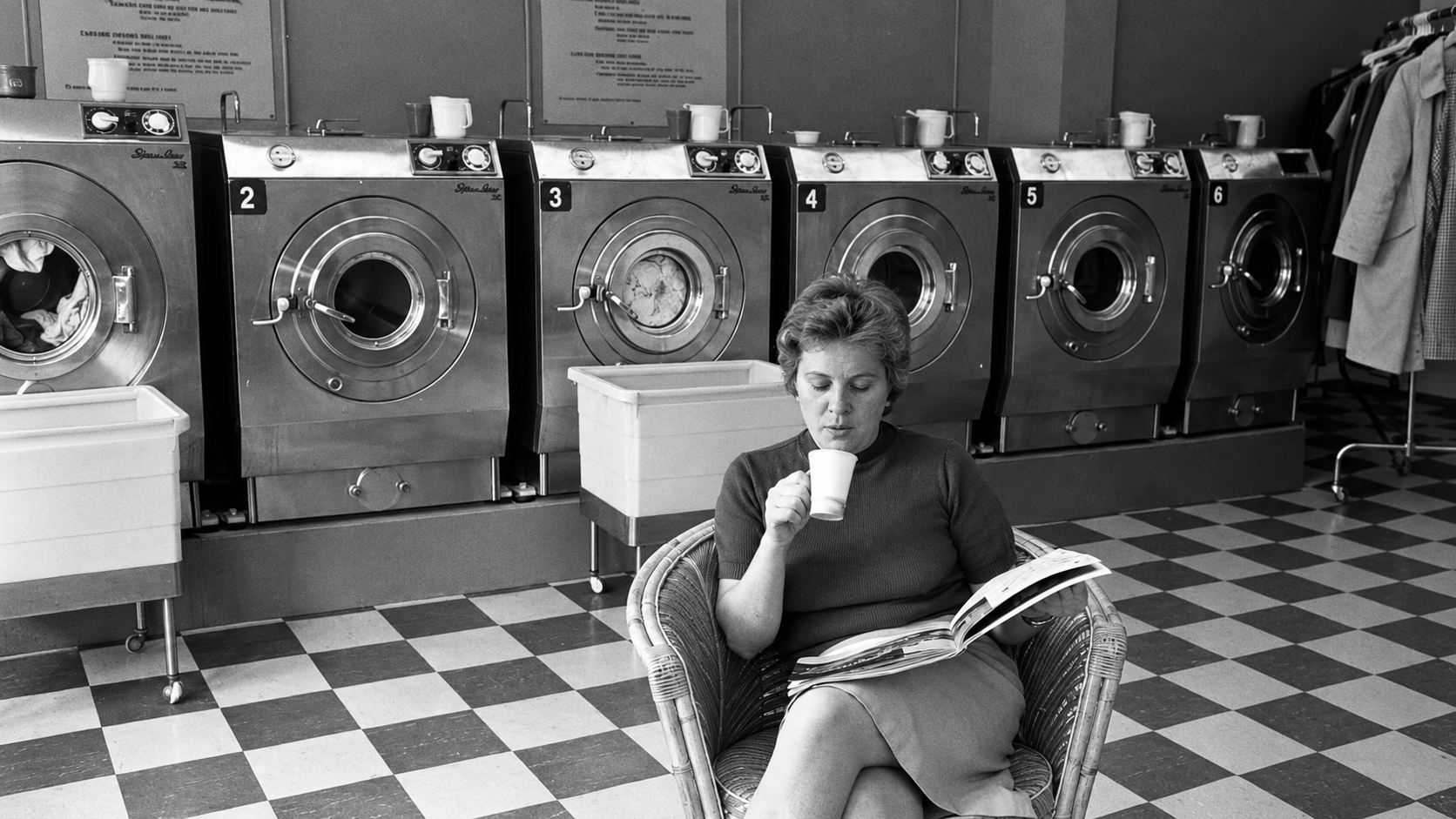 A woman sits in a chair at a laundromat, sipping from a cup and reading a magazine. Behind her are five front-loading washing machines and laundry baskets on a checkered floor.