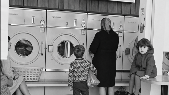 Four people sit and stand in front of large washing machines at a laundromat. A young boy stands with a woman, while a girl sits on a counter. Laundry baskets and a cardboard box are visible on the checkered floor.