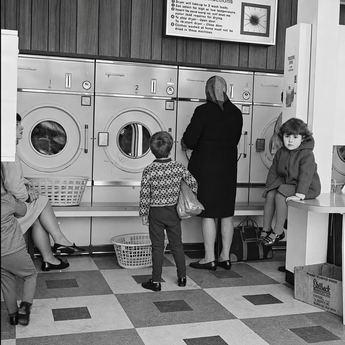 Four people sit and stand in front of large washing machines at a laundromat. A young boy stands with a woman, while a girl sits on a counter. Laundry baskets and a cardboard box are visible on the checkered floor.