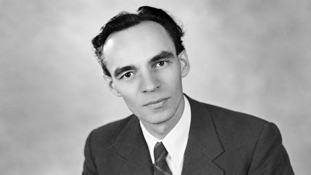 Black and white portrait of a man in a suit and tie, with dark hair, looking directly at the camera against a plain background.