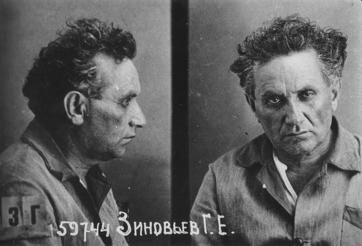 A black-and-white 1930s Soviet mugshot of Grigory Zinoviev, showing profile and frontal views. He is a middle-aged man with dark curly hair wearing a collared shirt. A number tag is on his shoulder, and his name is written in Russian below.